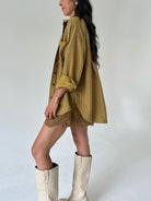 A woman wearing an oversized mustard yellow shirt with a lace hem shorts, styled with cream-colored knee-high boots against a plain light gray studio background.