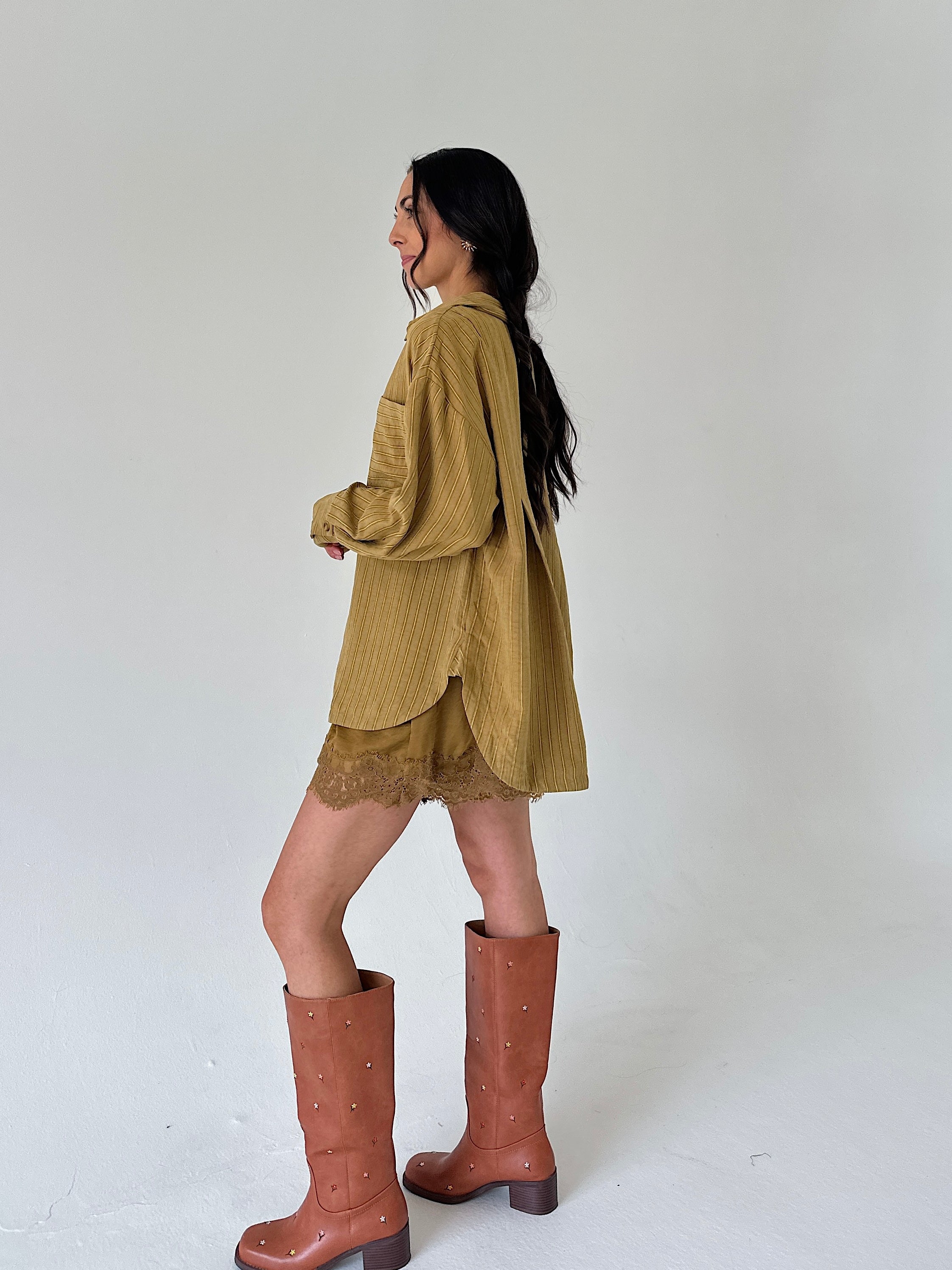 A woman is shown from the side wearing a mustard yellow shirt, matching lace hem shorts, and brown studded knee-high boots against a plain white studio background.