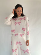 Woman wearing a white sweater and skirt with pink bow patterns against a plain background