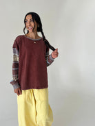 A woman modeling a rust-brown long-sleeve top with patterned knit sleeves and yellow cargo pants, standing against a plain off-white wall.
