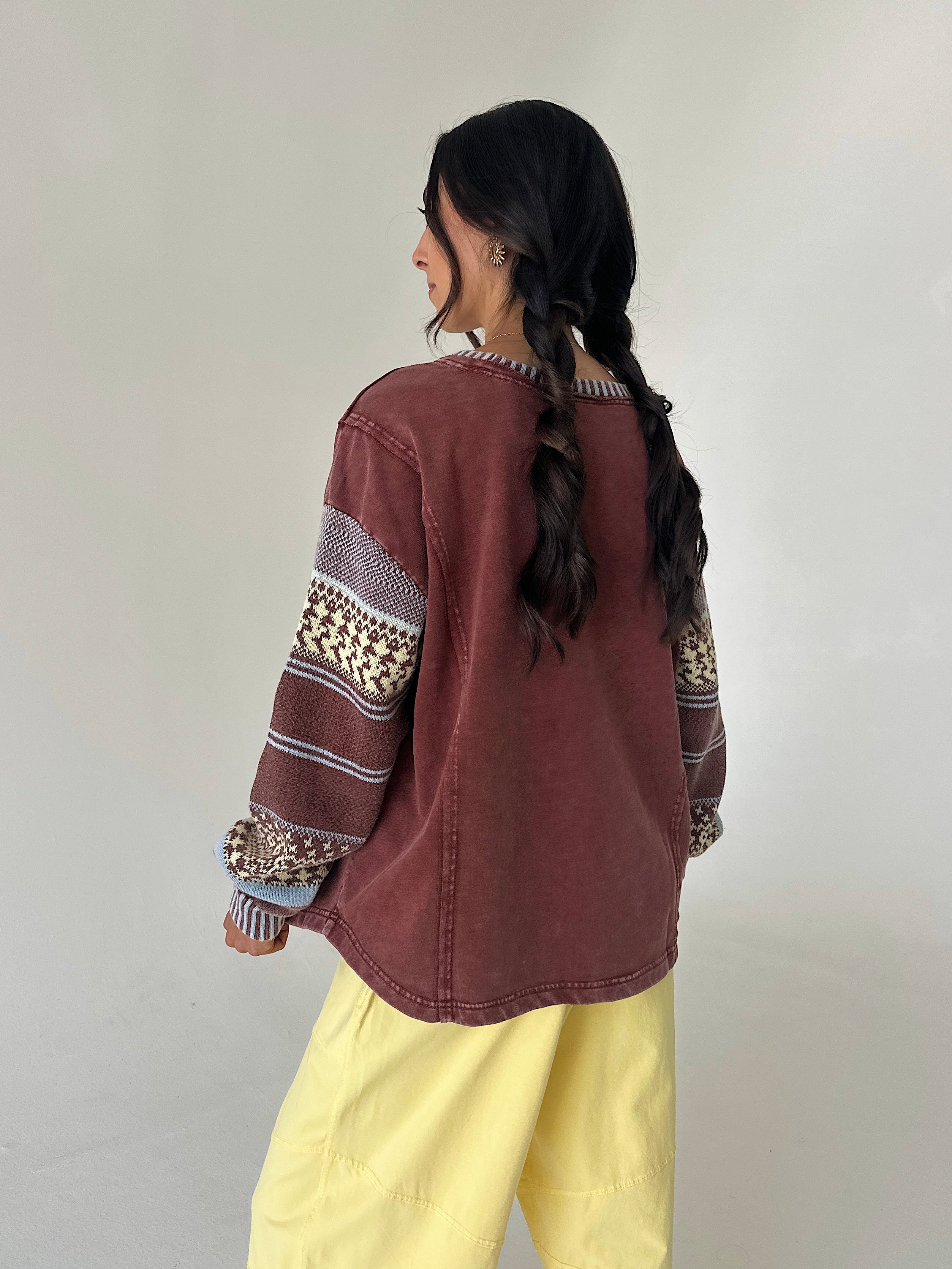 A woman with long dark hair in a braid is shown from the back, wearing a washed reddish-brown cropped sweatshirt with patterned knit sleeves and yellow pants, standing against a plain light gray wall.