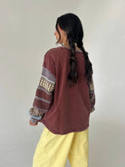 A woman with long dark hair in a braid is shown from the back, wearing a washed reddish-brown cropped sweatshirt with patterned knit sleeves and yellow pants, standing against a plain light gray wall.