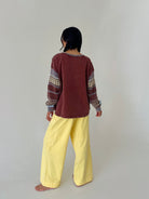 A person standing barefoot against a plain light gray studio background, shown from the back. They are wearing a loose long-sleeve top in dark reddish brown with patterned knit cuffs and shoulders, paired with wide-leg light yellow pants.