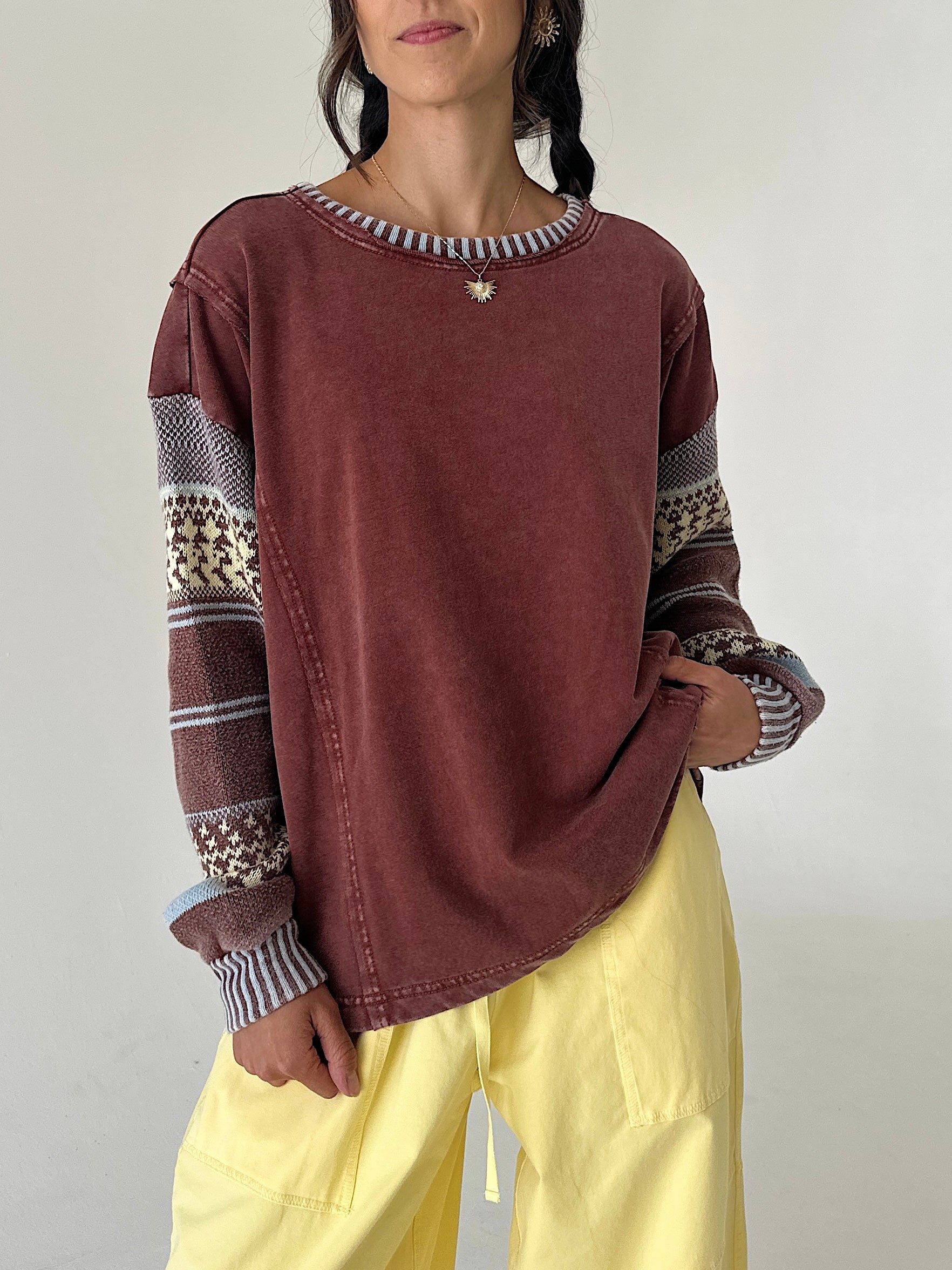 A woman wearing a washed dark reddish-brown long-sleeve sweatshirt with patterned knit cuffs and a striped crew neck, paired with light yellow pants, against a plain light gray background.