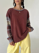 A woman wearing a washed dark reddish-brown long-sleeve sweatshirt with patterned knit cuffs and a striped crew neck, paired with light yellow pants, against a plain light gray background.