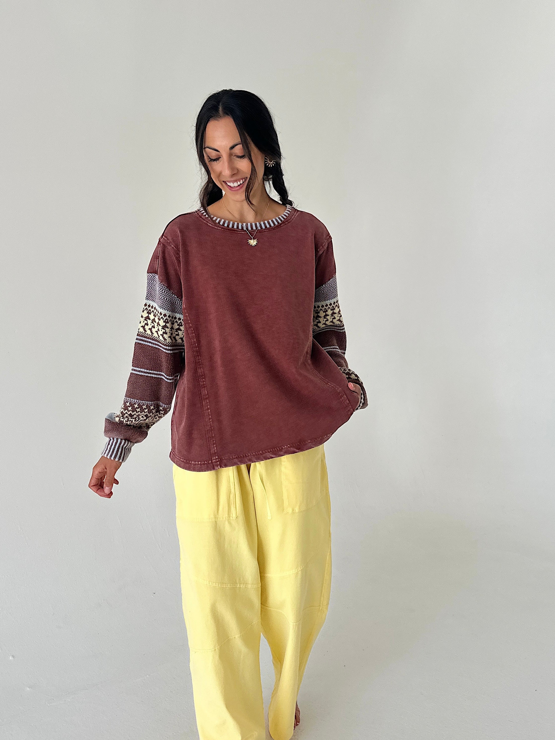 A woman modeling a rust-brown long-sleeve top with patterned gray and cream sleeves, paired with loose yellow pants, against a plain light gray studio background.