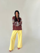A woman modeling a bohemian-style outfit consisting of a rust-brown long-sleeve top with patterned sleeves and bright yellow wide-leg pants, standing against a plain off-white studio background.