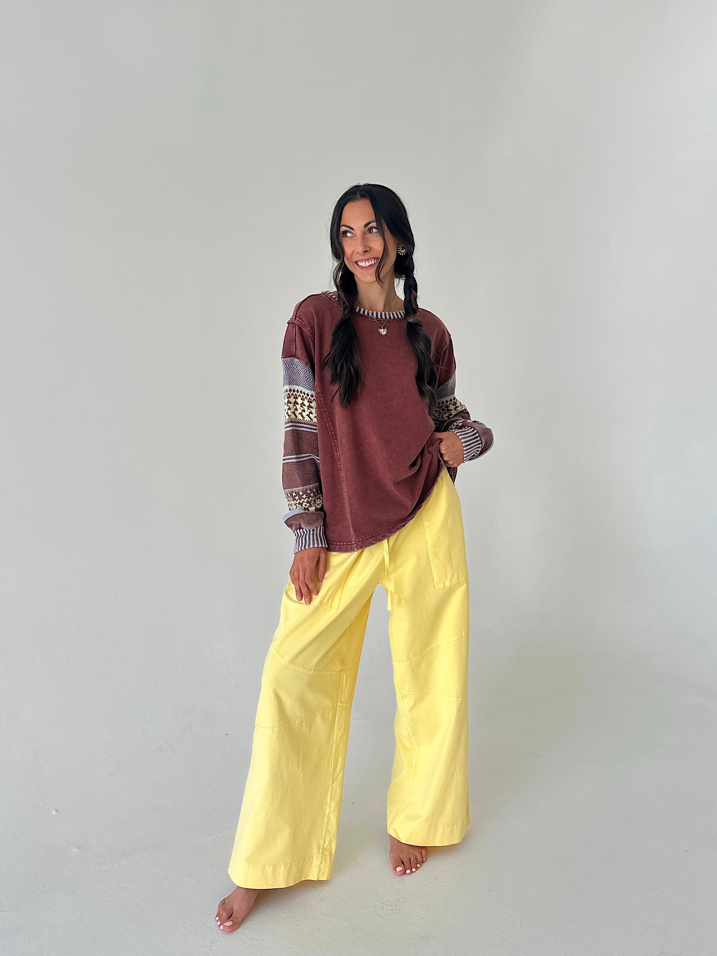 A woman modeling a two-piece outfit consisting of a rust-brown and gray patterned long-sleeve top and bright yellow wide-leg pants, standing against a plain light gray studio background.
