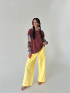A woman modeling a two-piece outfit consisting of a rust-brown and gray patterned long-sleeve top and bright yellow wide-leg pants, standing against a plain light gray studio background.