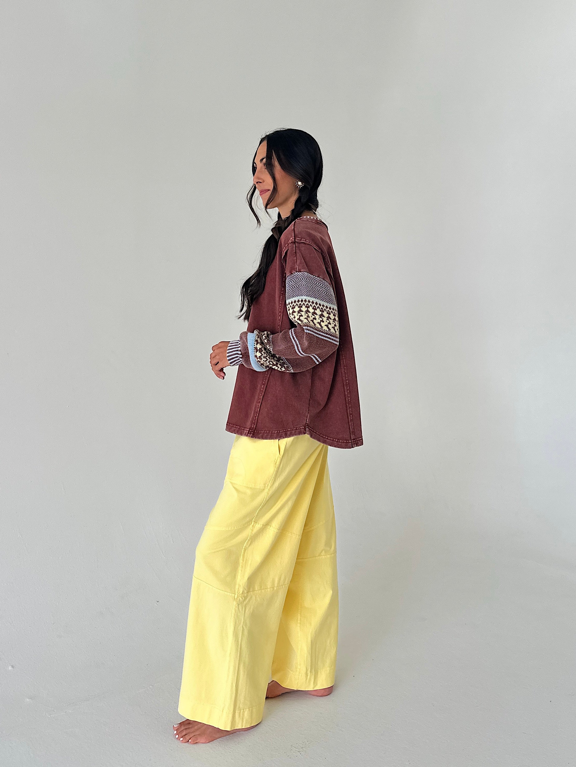 A woman stands barefoot in profile wearing a rust-brown cropped jacket with patterned knit sleeves and bright yellow wide-leg pants against a plain light gray studio background.