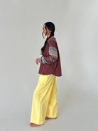 A woman stands barefoot in profile wearing a rust-brown cropped jacket with patterned knit sleeves and bright yellow wide-leg pants against a plain light gray studio background.