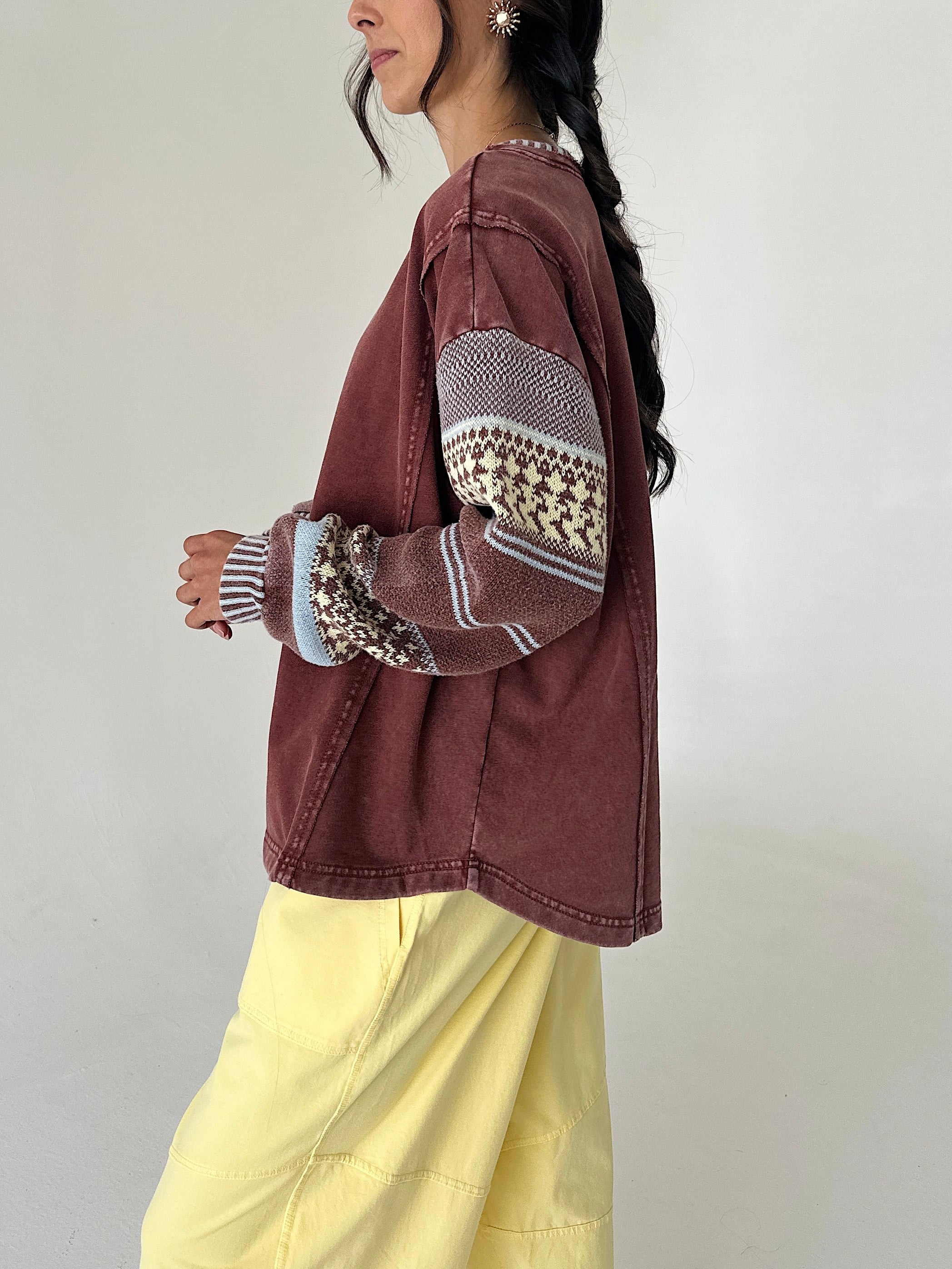 A woman wearing a washed brownish-red cropped jacket with patterned knit sleeves, paired with light yellow pants, shown from a side profile against a plain light gray wall.