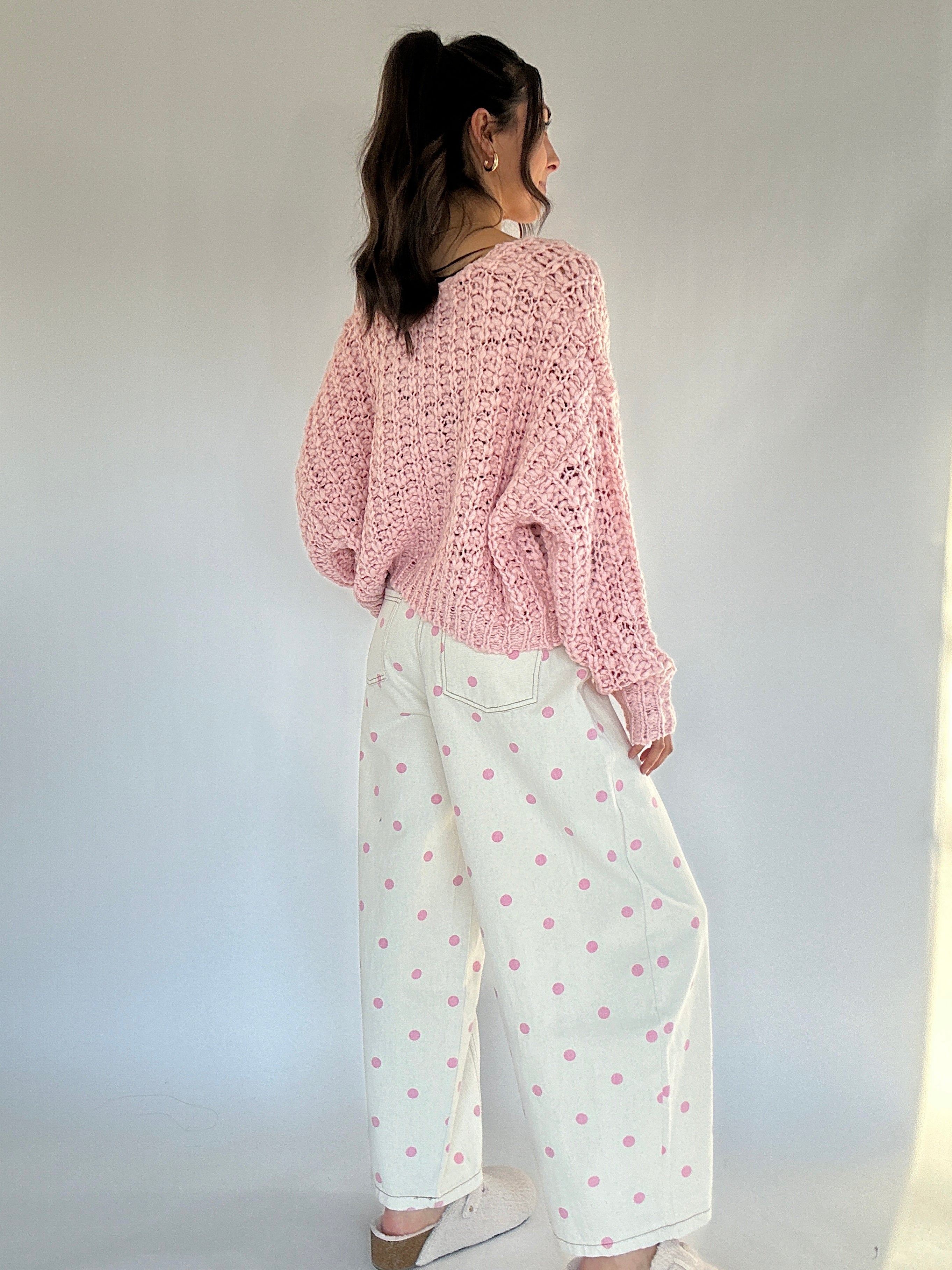 Woman wearing a pink knitted sweater and white pants with pink polka dots against a plain background showing back