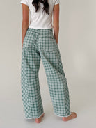 A person shown from the back wearing green and white plaid wide-leg pants, a white t-shirt, and barefoot on a light neutral studio background.