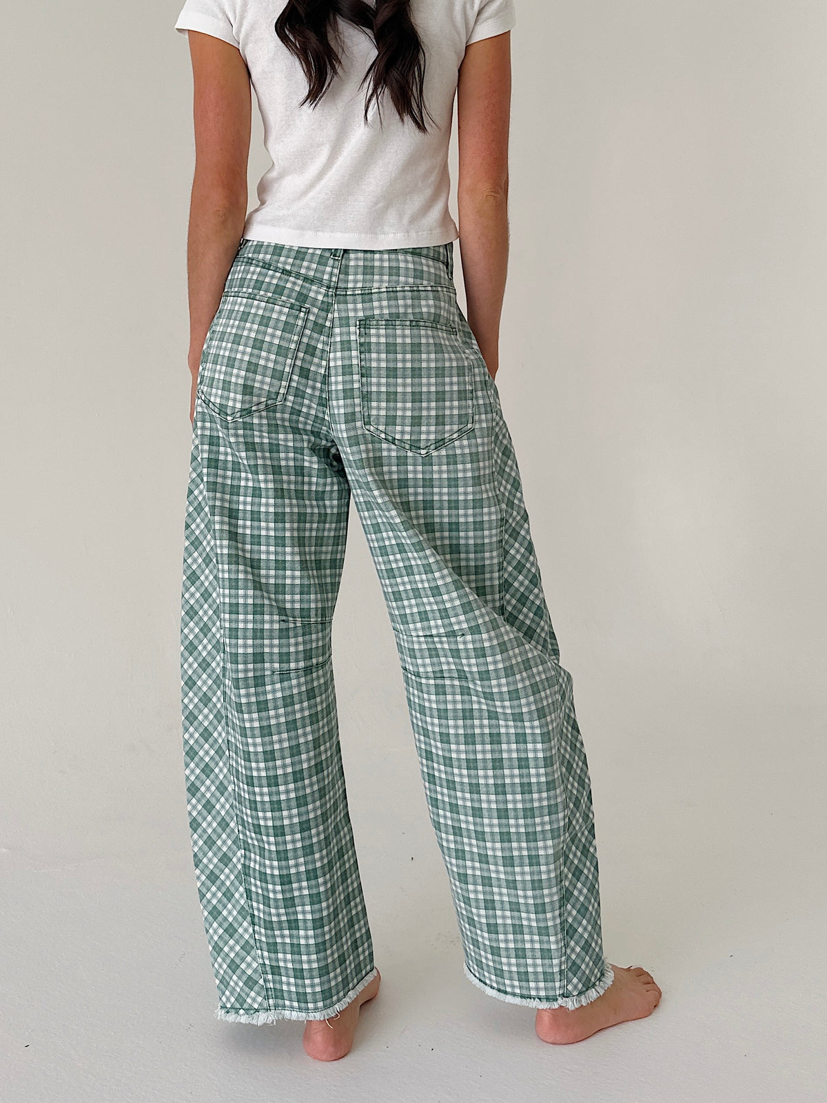 A person shown from the back wearing green and white plaid wide-leg pants, a white t-shirt, and barefoot on a light neutral studio background.
