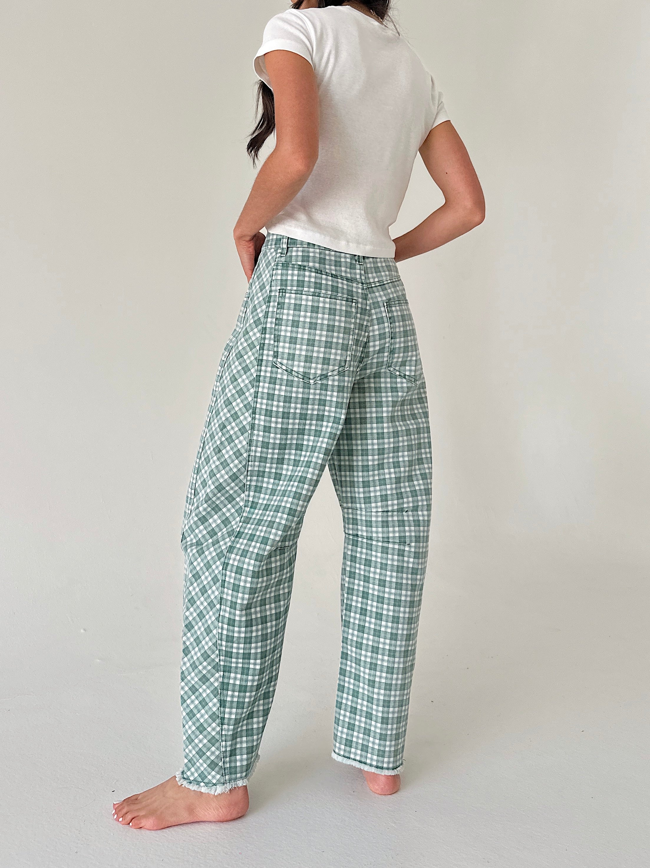 A woman shown from the back wearing green and white plaid pants, a white t-shirt, and standing barefoot against a plain light gray studio background.