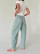 A woman shown from the back wearing green and white plaid pants, a white t-shirt, and standing barefoot against a plain light gray studio background.
