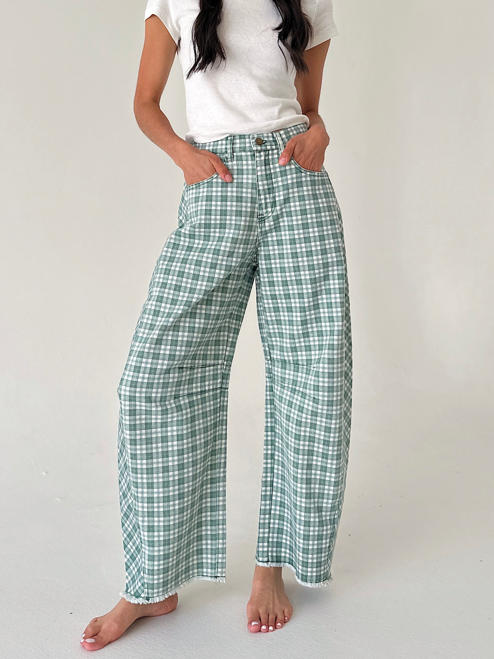 A model wearing green and white plaid wide-leg pants with a white t-shirt, standing barefoot against a light gray studio background.