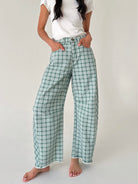 A model wearing green and white plaid wide-leg pants with a white t-shirt, standing barefoot against a light gray studio background.
