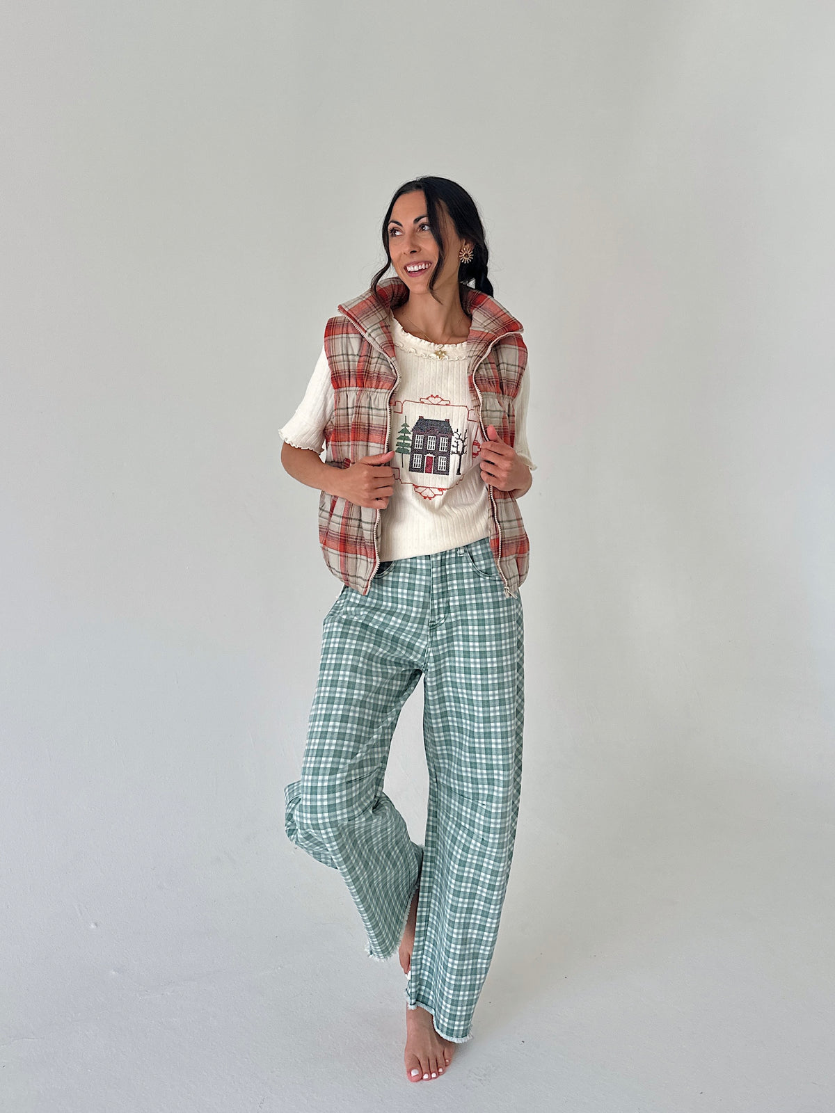 A woman stands barefoot against a plain light gray studio background, wearing a cream graphic T-shirt, a red and green plaid vest, and green-and-white plaid pants.