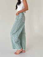 A woman wearing green and white plaid wide-leg pants with a white sleeveless top, standing barefoot against a plain light gray studio background.
