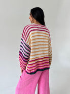 A woman shown from the back wearing a chunky knit striped sweater with horizontal bands of pink, cream, tan, and black, paired with bright pink pants, against a plain light gray background.