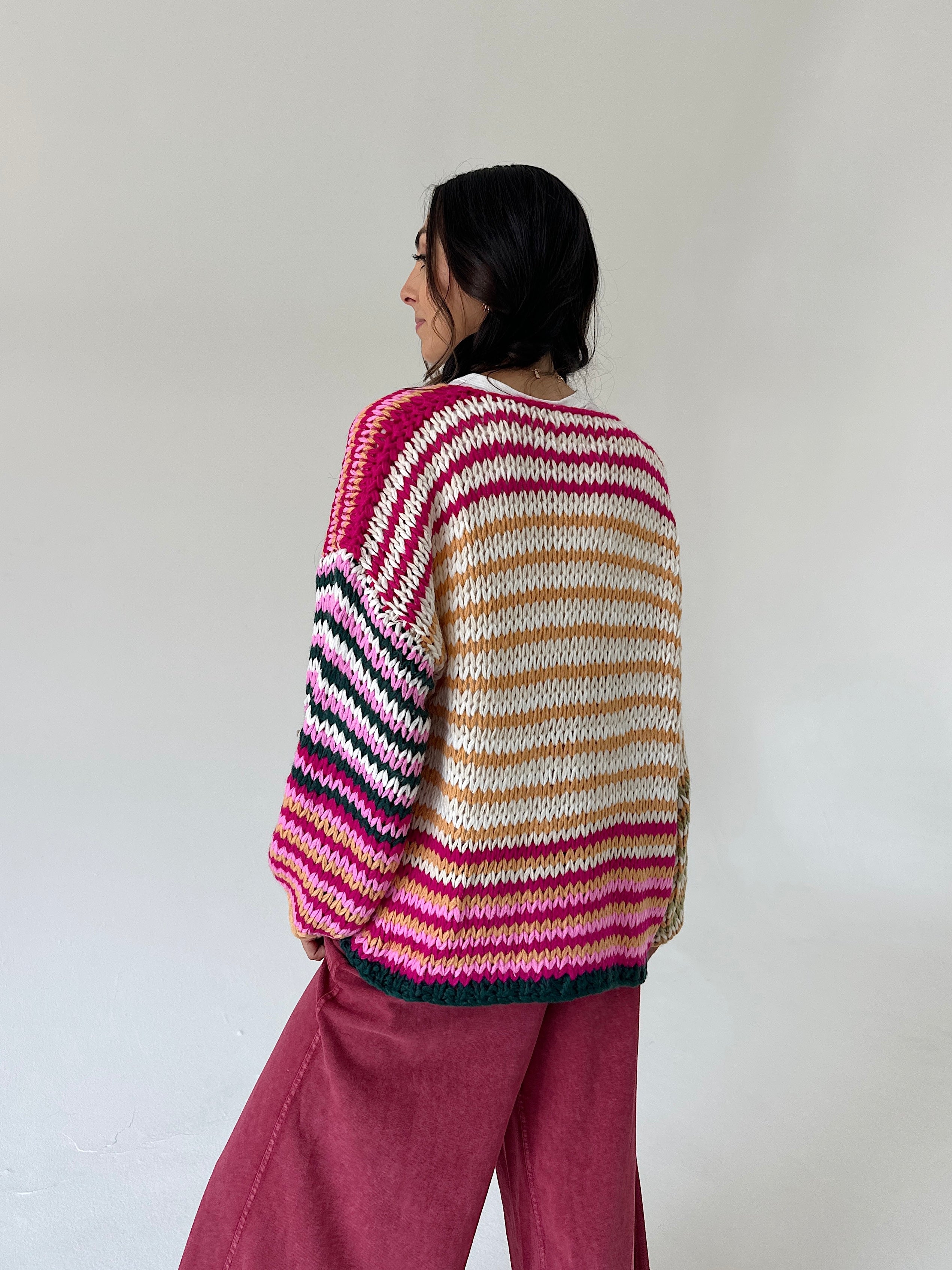 A woman is shown from behind wearing a chunky, hand-knit striped sweater with horizontal bands of pink, cream, tan, and dark green, paired with dusty rose wide-leg pants, set against a plain off-white wall.