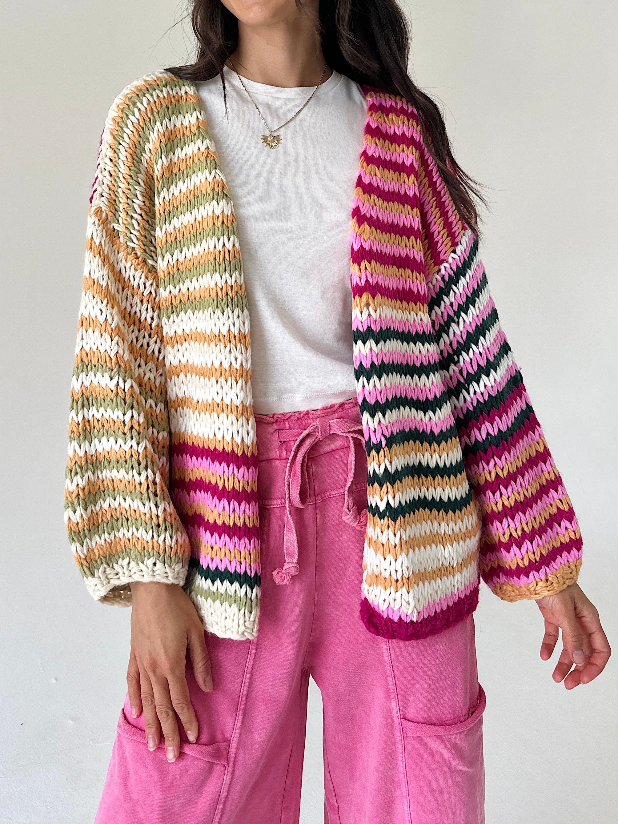 A person wearing a chunky knit open-front cardigan with horizontal multicolor stripes in cream, orange, green, pink, and black, layered over a white top and bright pink drawstring pants.