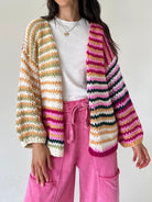 A person wearing a chunky knit open-front cardigan with horizontal multicolor stripes in cream, orange, green, pink, and black, layered over a white top and bright pink drawstring pants.