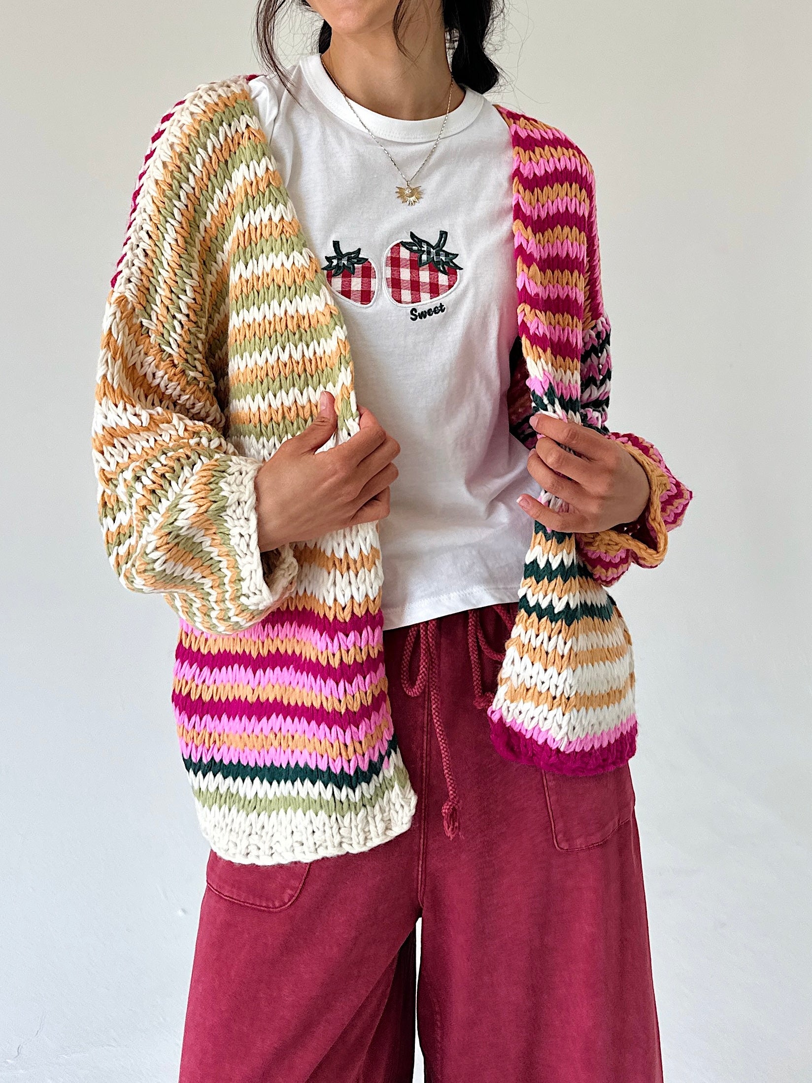 A person wearing a chunky hand-knit striped cardigan with cream, pink, orange, green, and purple stripes over a white t-shirt and burgundy pants, photographed against a plain off-white wall.