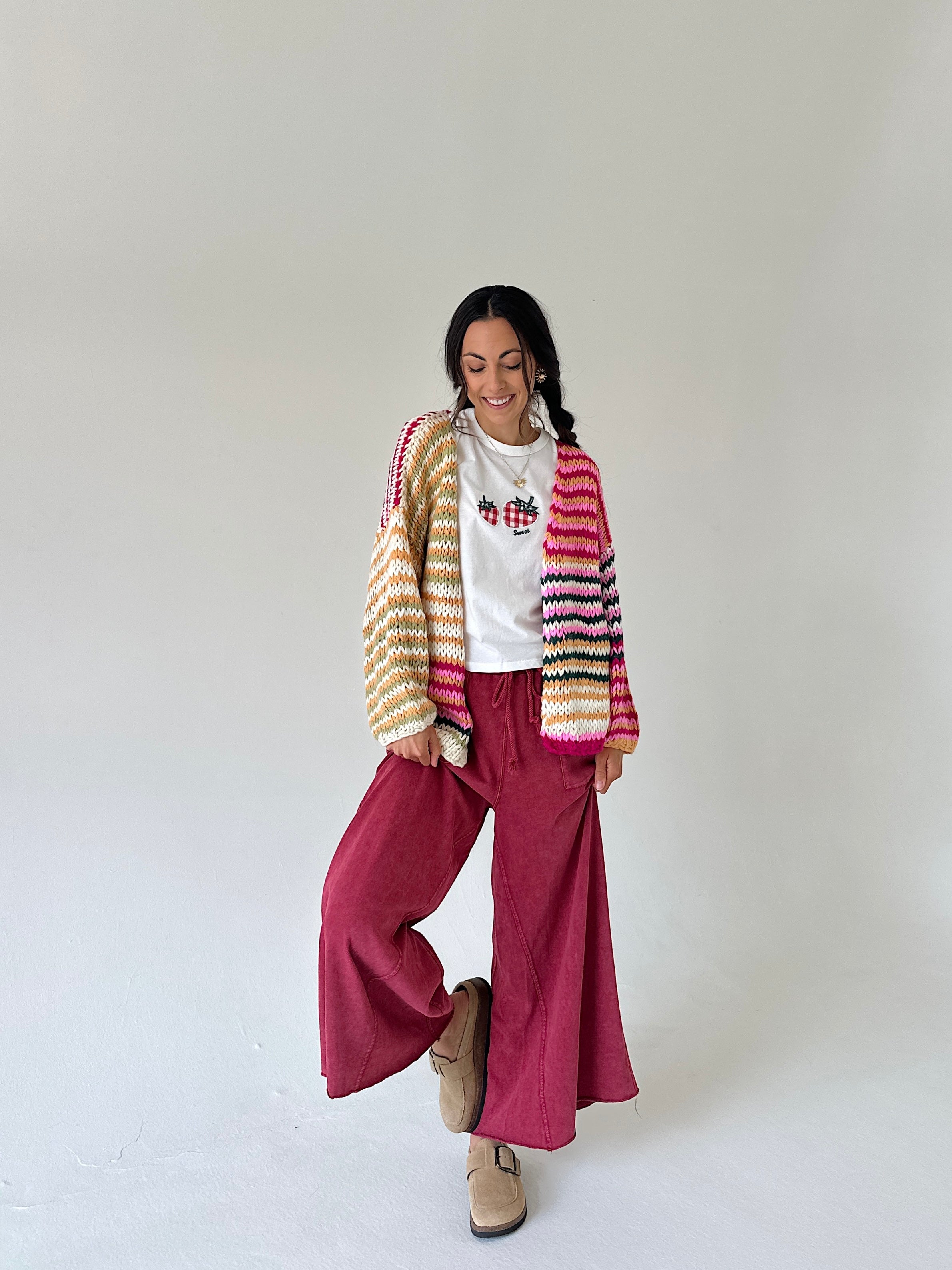 A woman modeling a colorful striped knit cardigan over a white t-shirt, paired with wide-leg burgundy pants and beige loafers, against a plain light gray studio background.