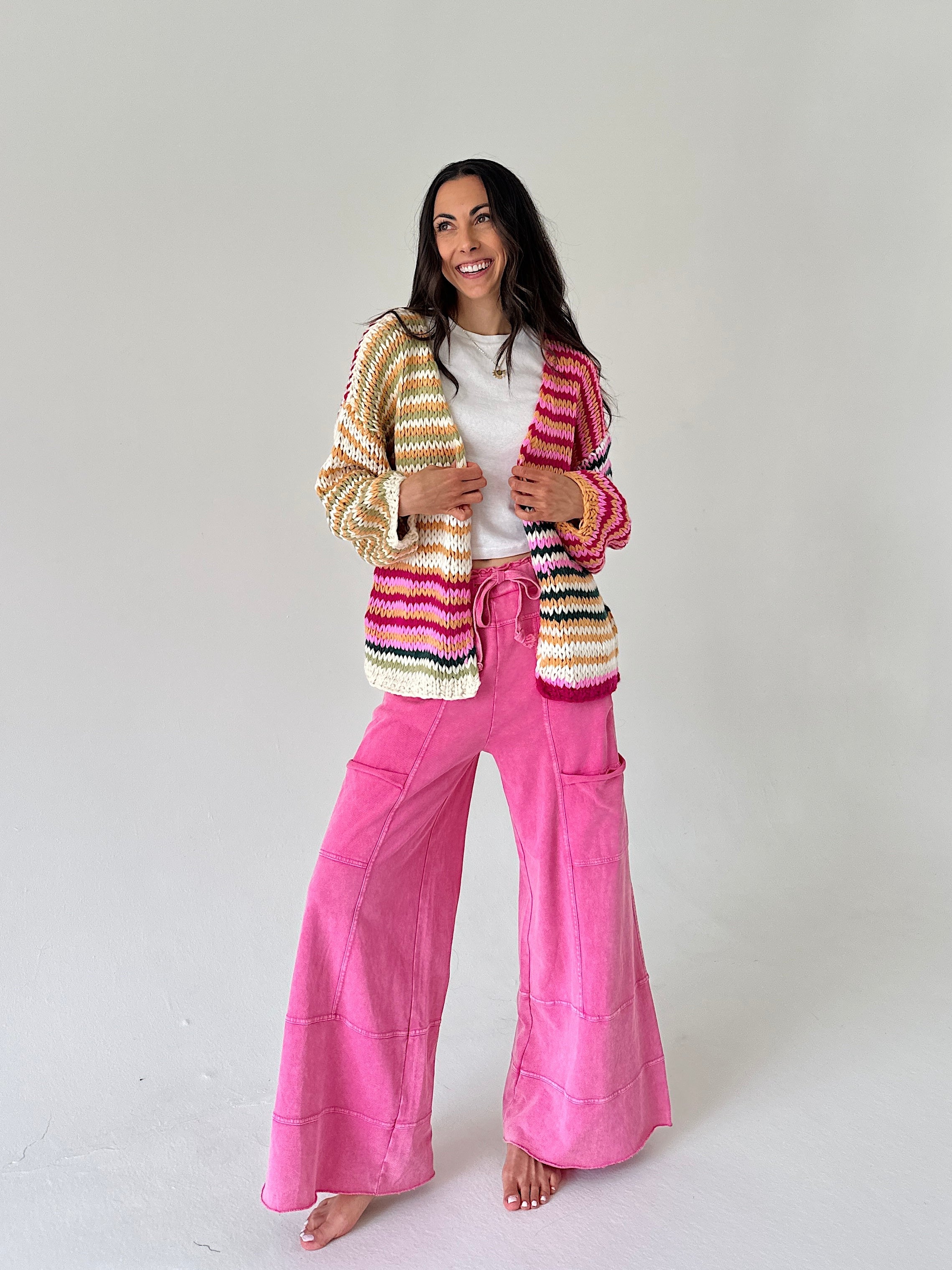 A woman stands barefoot against a plain light gray studio background, wearing a colorful striped knit cardigan over a white top and bright pink wide-leg pants.