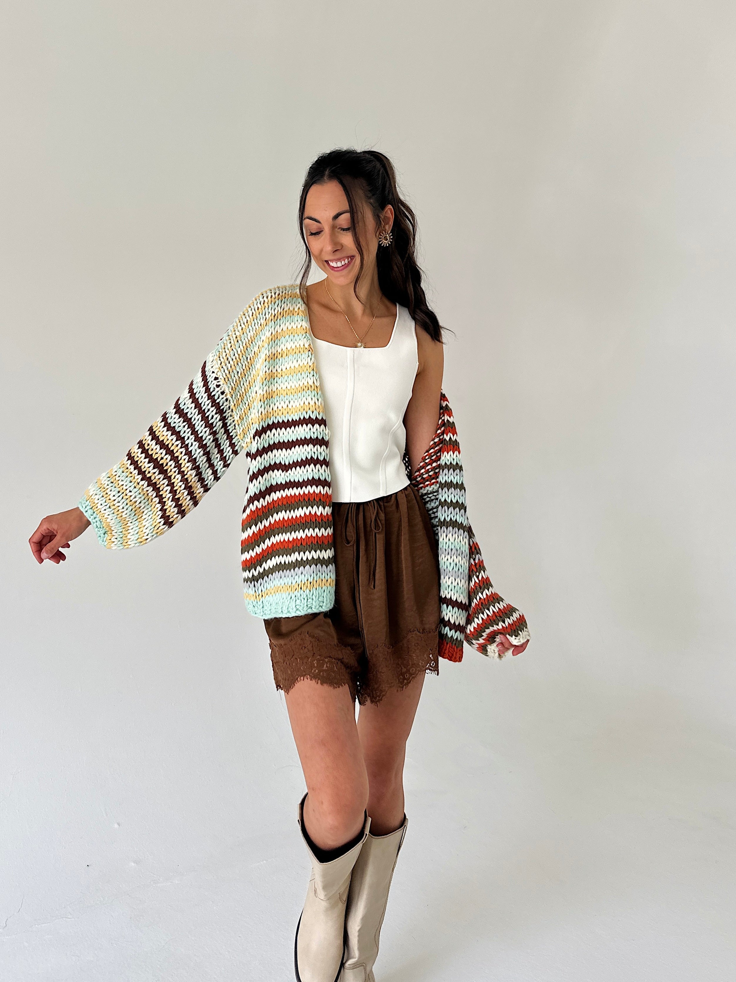 A woman modeling a chunky knit cardigan with horizontal stripes in cream, yellow, brown, orange, and light blue, worn over a white top and brown shorts.