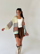 A woman modeling a chunky knit cardigan with horizontal stripes in cream, yellow, brown, orange, and light blue, worn over a white top and brown shorts.