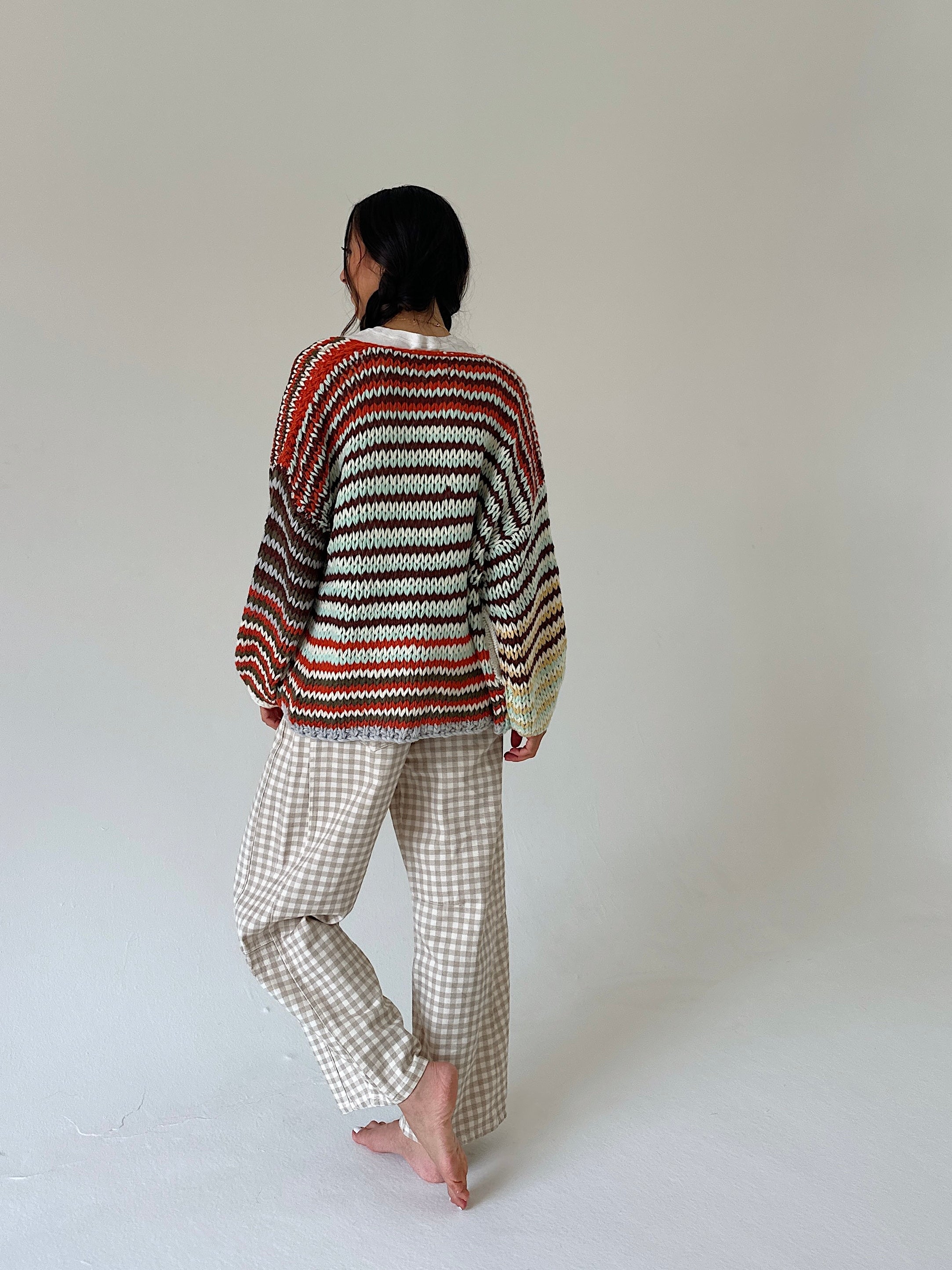 A person standing barefoot against a plain light gray studio background, viewed from the back. They are wearing a loose striped knit sweater with horizontal bands of rust red, cream, brown, and black, paired with light beige gingham pants.