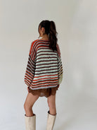 A woman is shown from behind wearing a chunky knit sweater with horizontal stripes in rust orange, brown, cream, and dark brown, paired with brown shorts and off-white boots, against a plain light gray background.