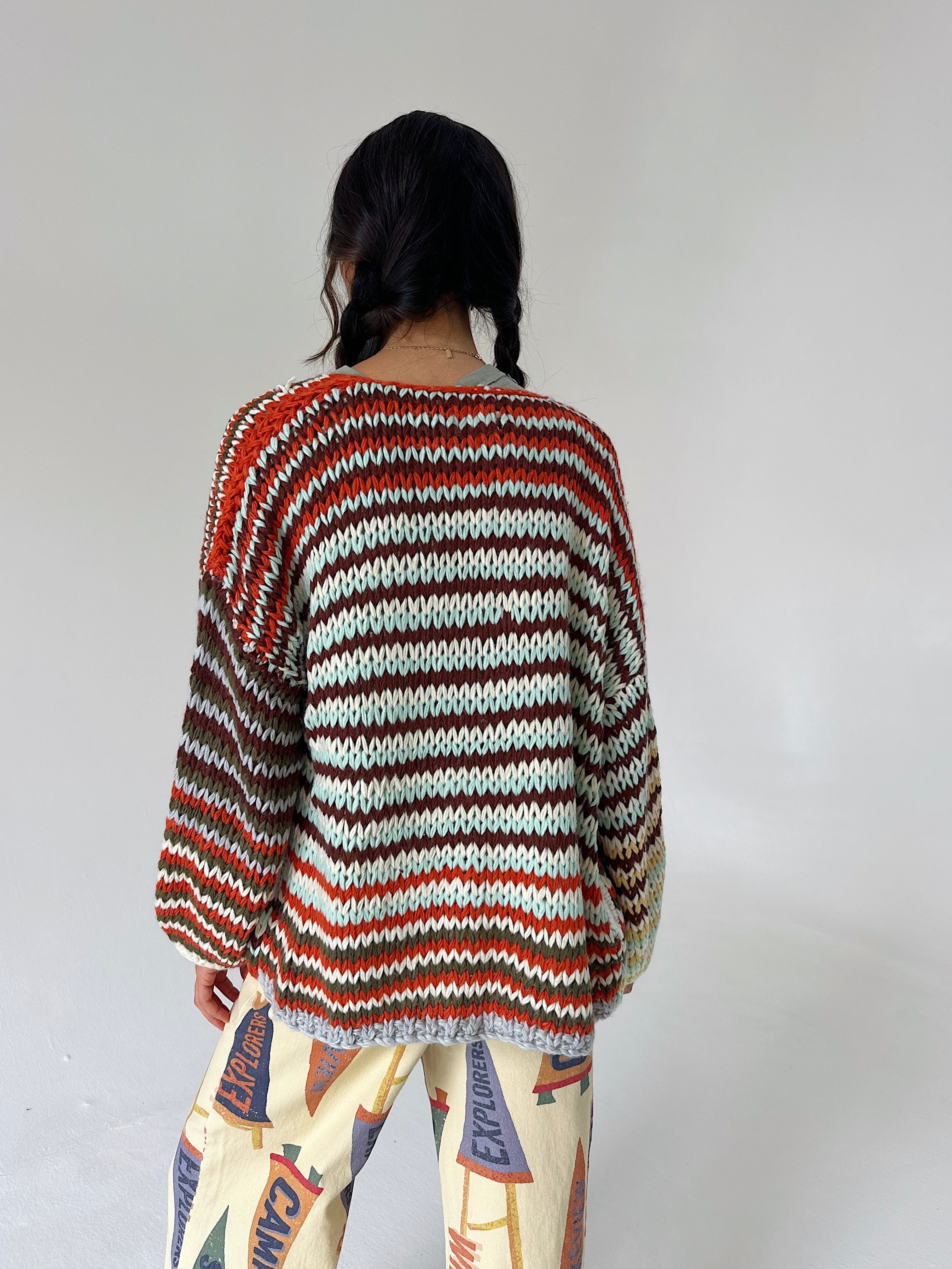 A person shown from the back wearing a loose, long-sleeve striped knit sweater with horizontal bands of rust red, dark brown, cream, and gray, paired with patterned pants against a plain light gray studio background.