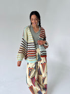 A woman modeling a chunky knit cardigan with horizontal multicolor stripes in beige, brown, red, and light blue, worn over a gray top and paired with cream wide-leg pants featuring a red, blue, and brown flag pattern.