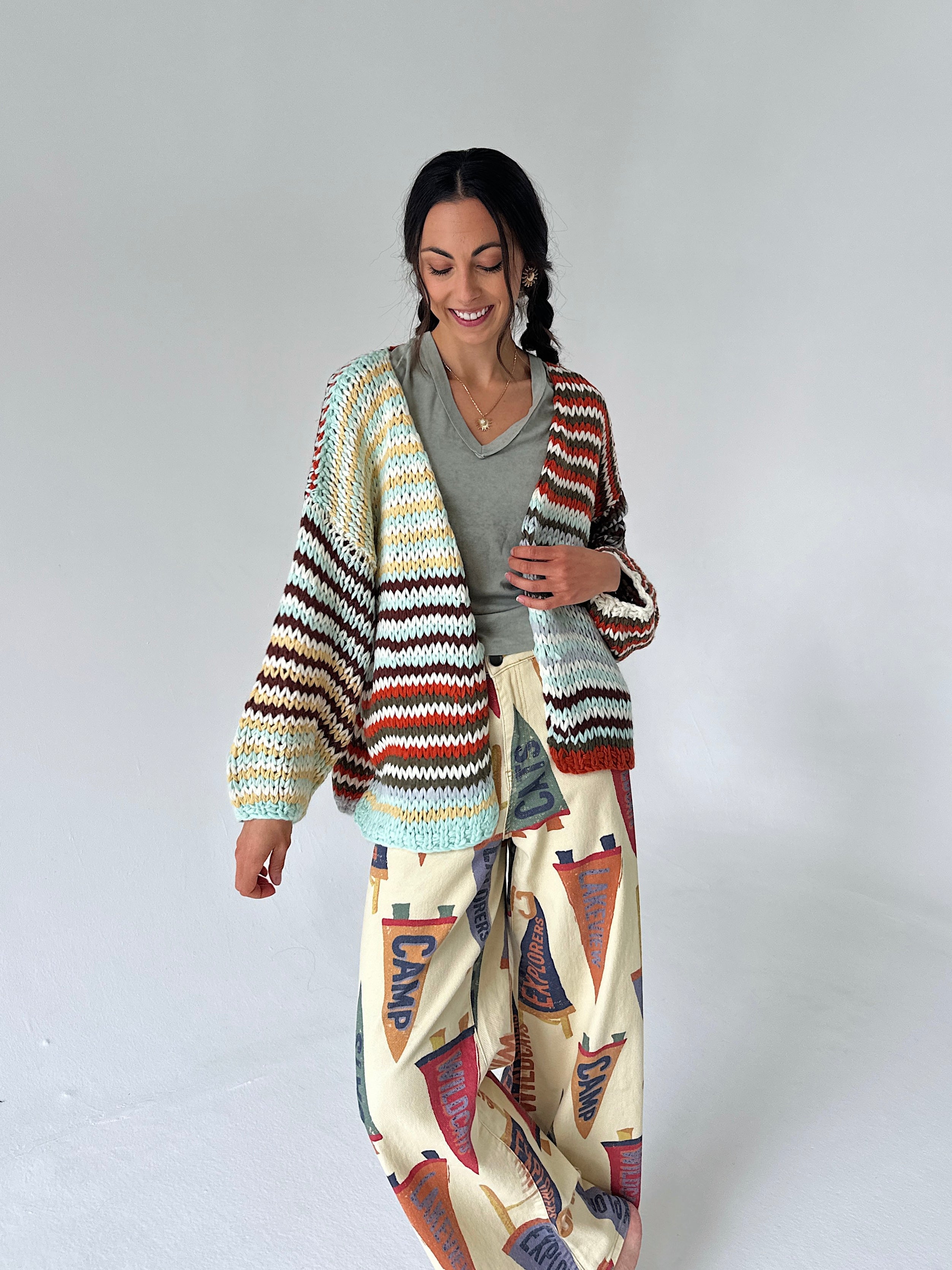A woman modeling a chunky knit cardigan with horizontal multicolor stripes in beige, brown, red, and light blue, worn over a gray top and paired with cream wide-leg pants featuring a red, blue, and brown flag pattern.