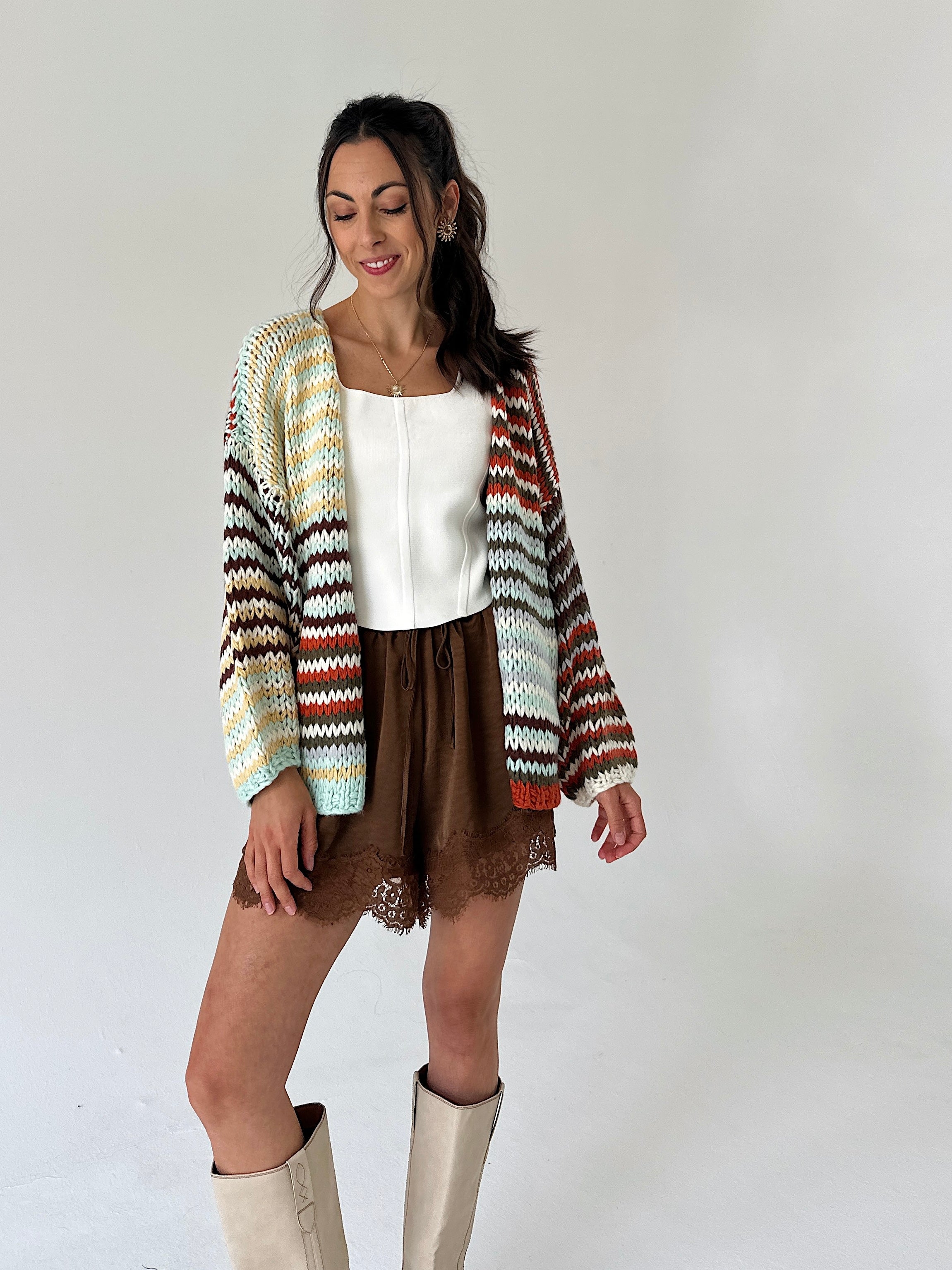 A woman modeling a long-sleeve knit cardigan with a multicolor chevron pattern in cream, orange, brown, and green, worn over a white top and brown shorts.