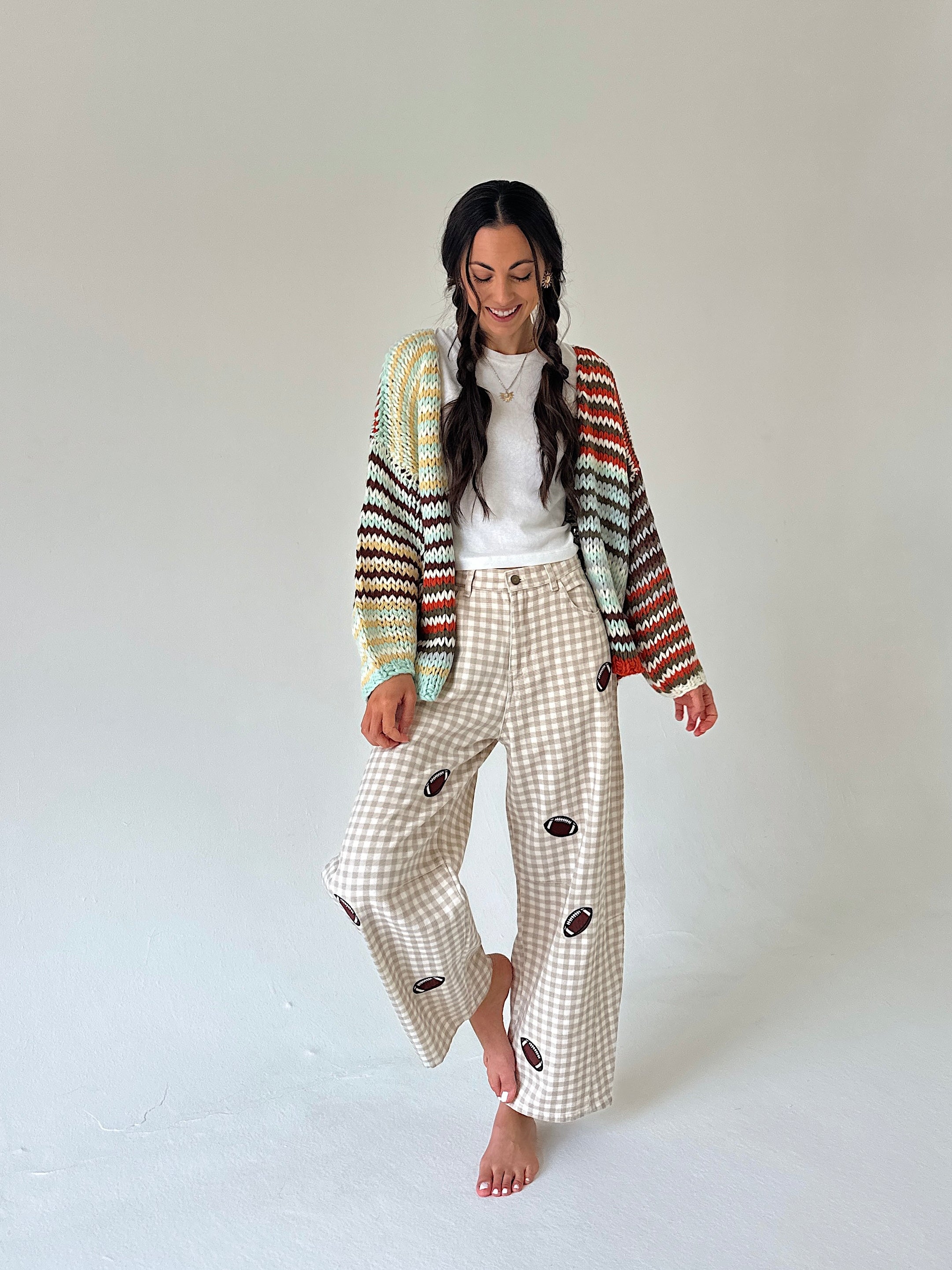 A woman stands barefoot against a plain light gray studio background, wearing a cream gingham wide-leg pant set with dark brown football patches, a white top, and a multicolor striped knit cardigan.