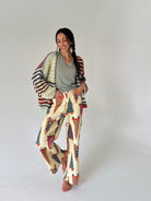 A woman modeling a bohemian-style outfit consisting of wide-leg cream pants with a colorful penant print, a gray V-neck t-shirt, and a striped knit cardigan draped over her shoulders, set against a plain light gray studio background.