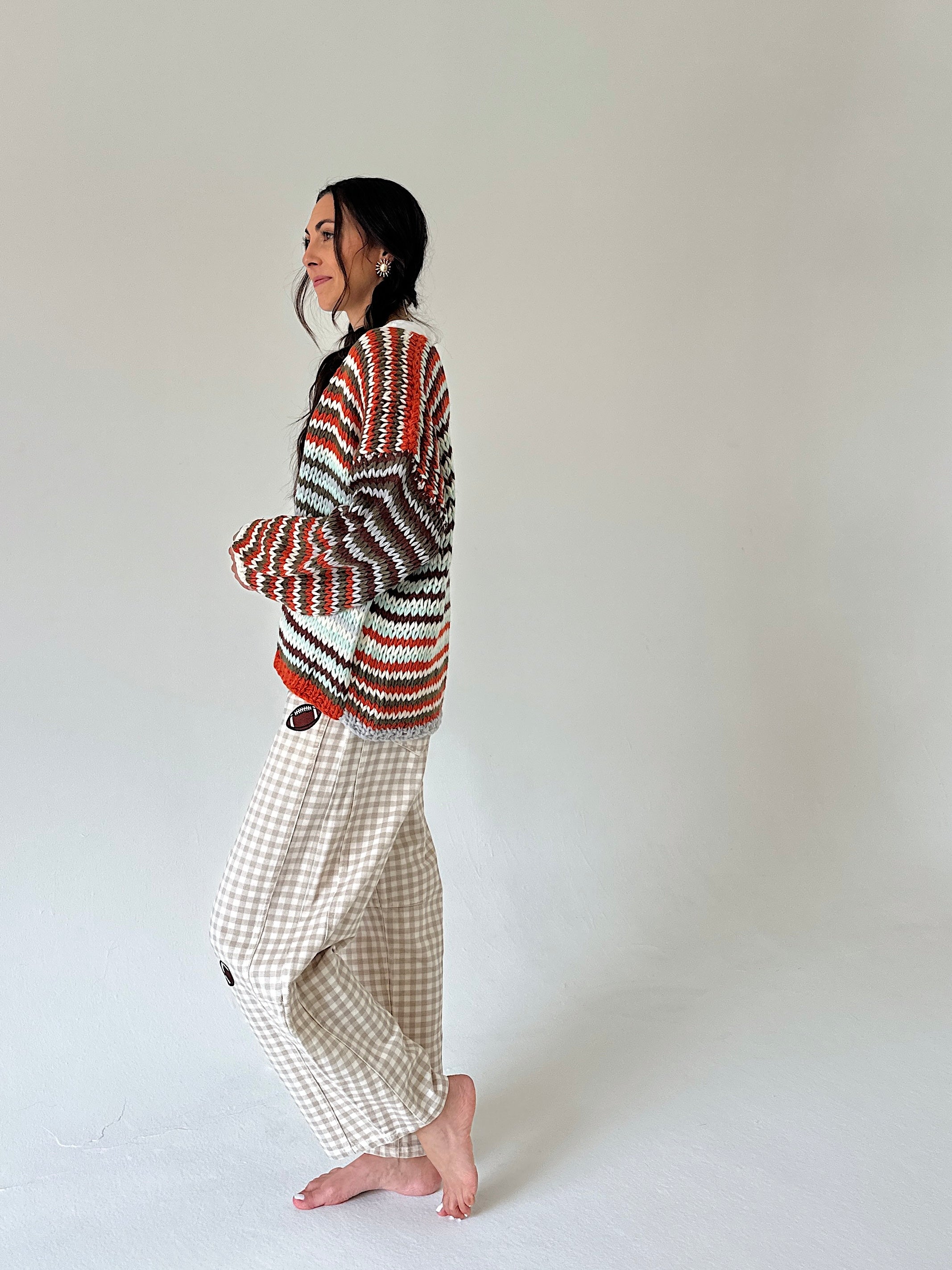 A woman stands barefoot in profile against a plain light gray studio background, wearing a multicolor knit sweater with red, orange, black, and white stripes and beige gingham pants.