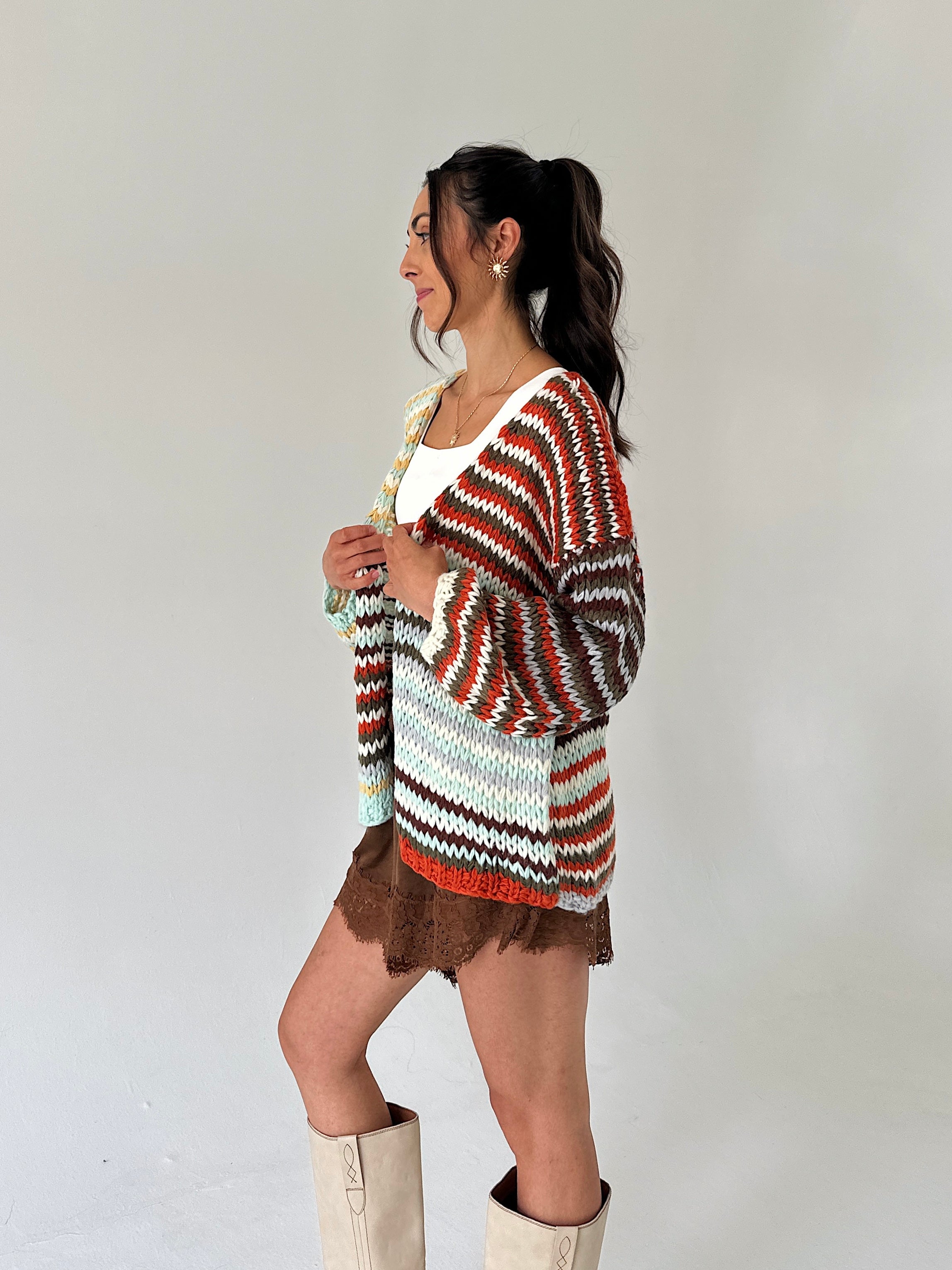 A woman modeling a chunky knit cardigan with horizontal wavy stripes in rust orange, brown, white, and mint green, styled with a white top, brown skirt, and beige boots against a plain light gray background.