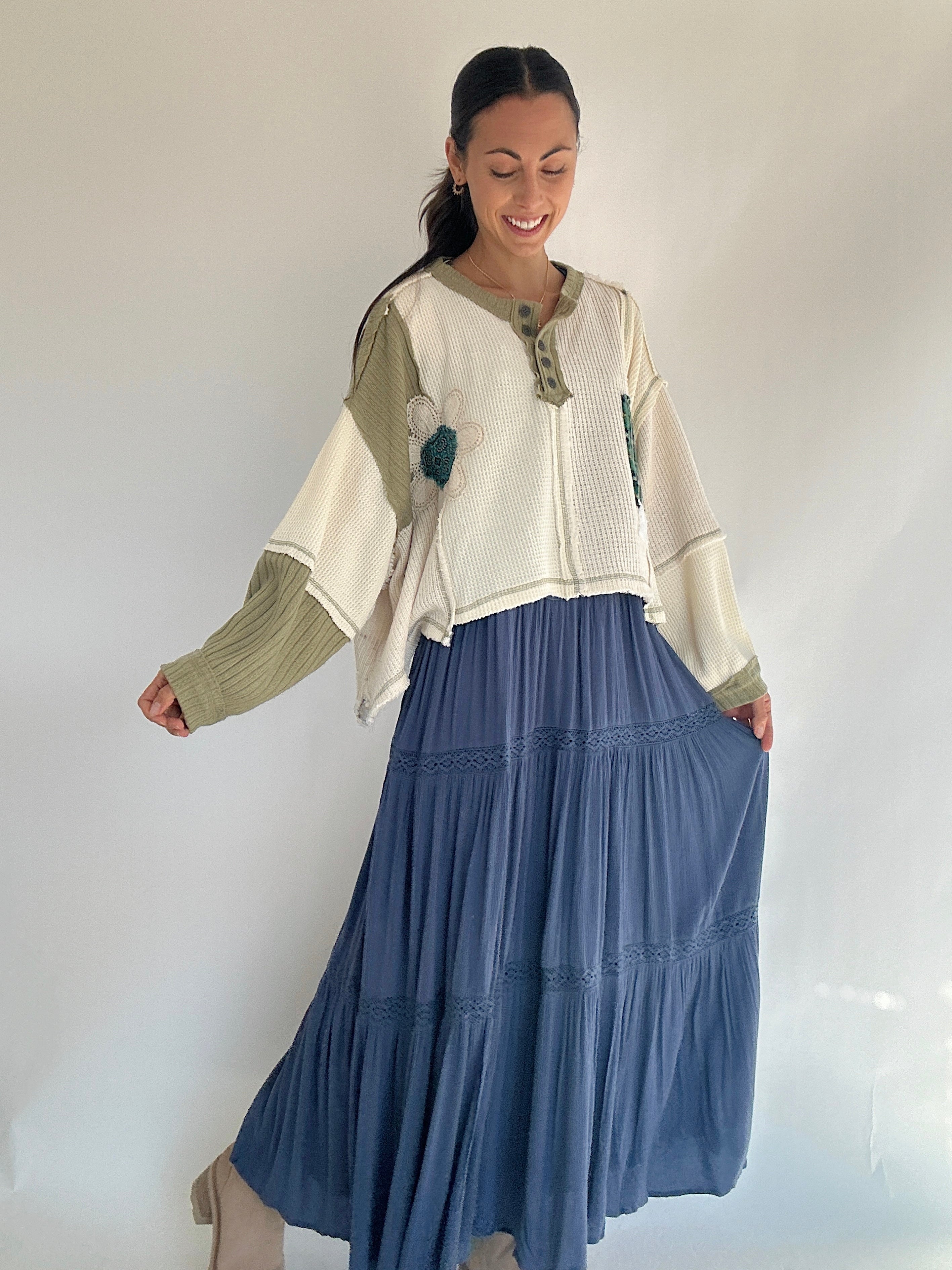 Woman wearing a layered outfit with a white and green knit tip over a blue overall dress making it look like a skirt against a plain background