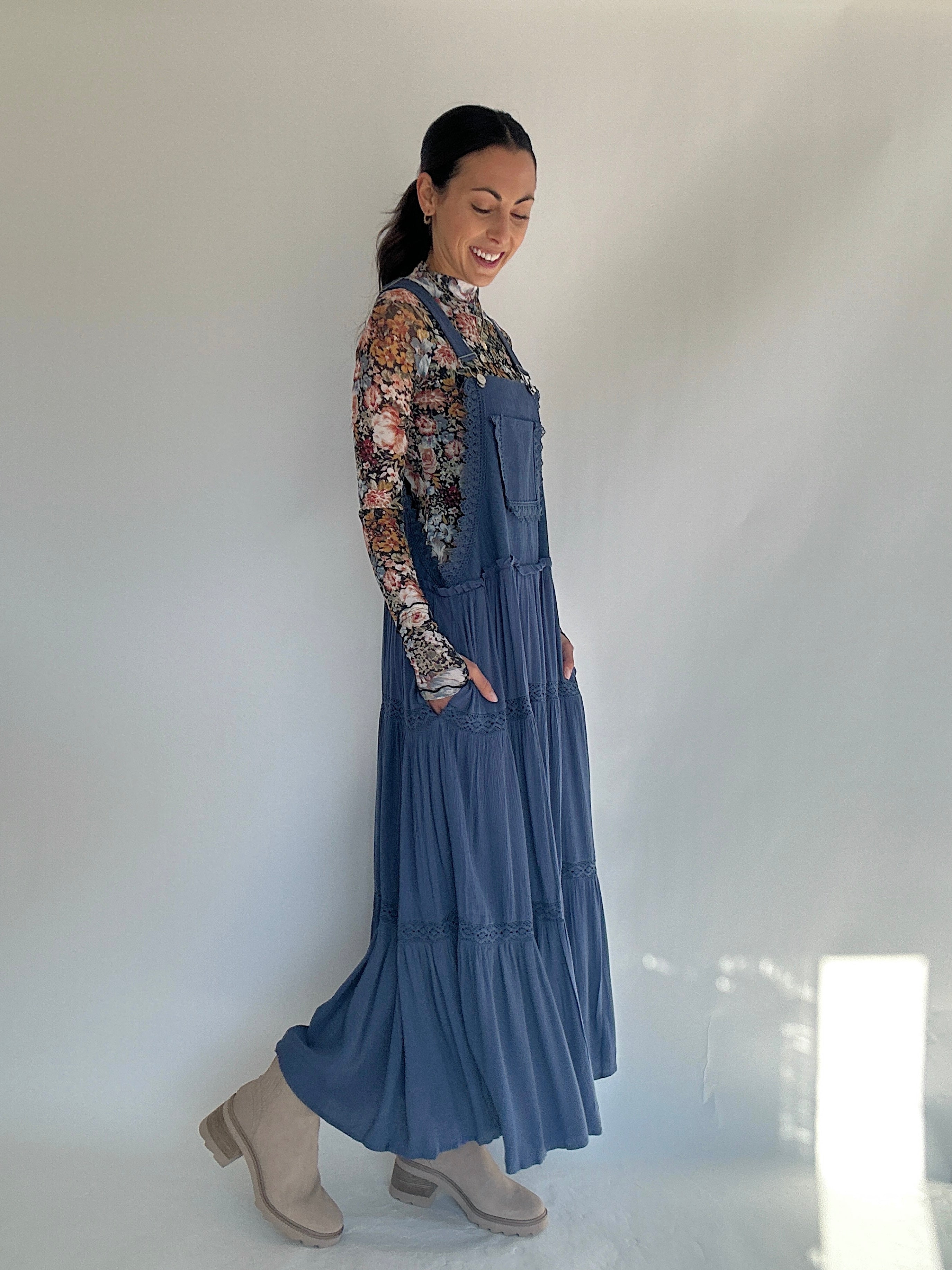 Woman wearing a long blue denim dress with a floral blouse against a white wall showing side view and pockets