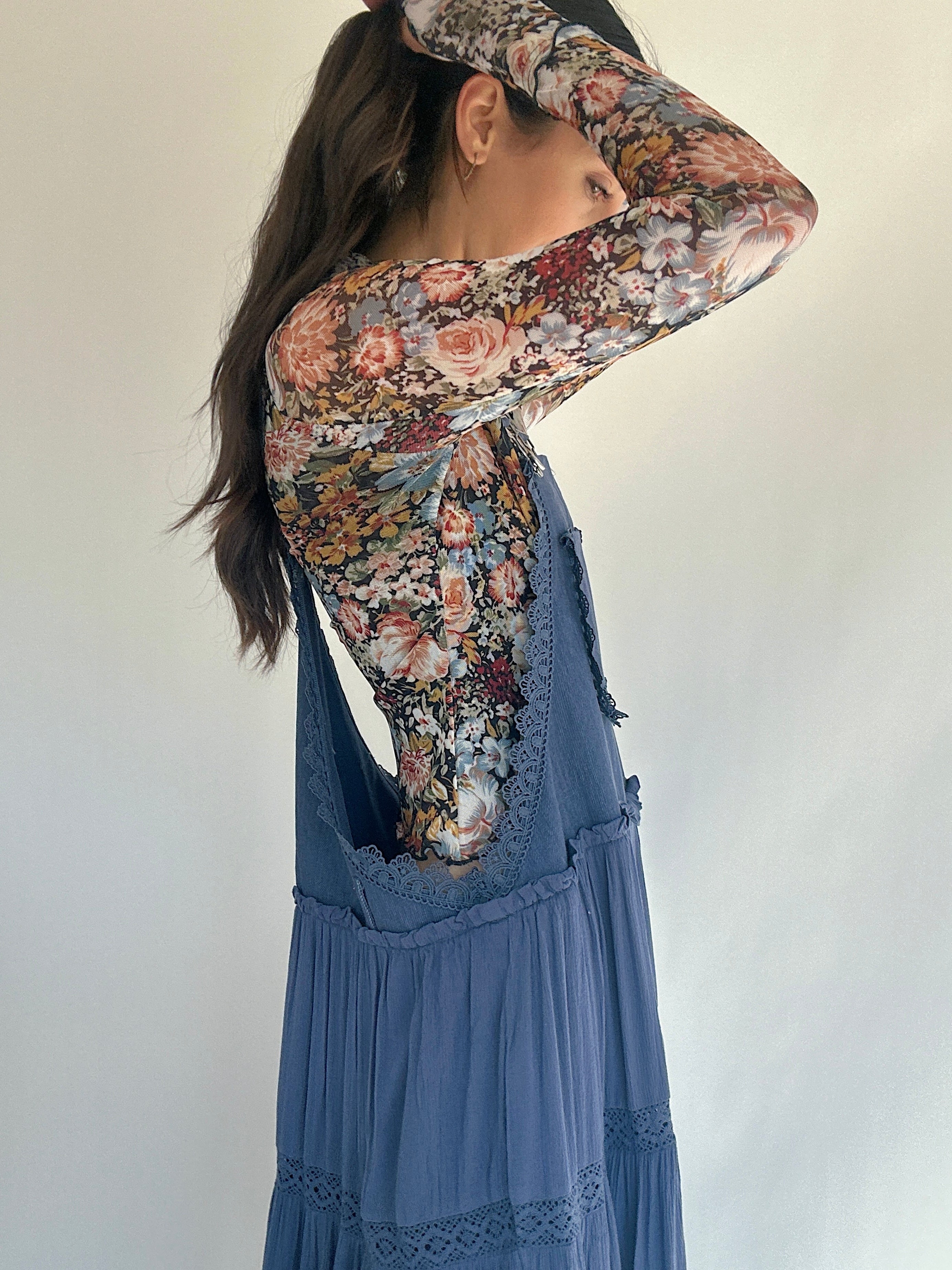 Woman wearing a floral top under a blue overall dress against a plain background to show off side view