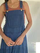 Top of a blue denim overall dress with lace details and front pocket worn by a person on a plain background