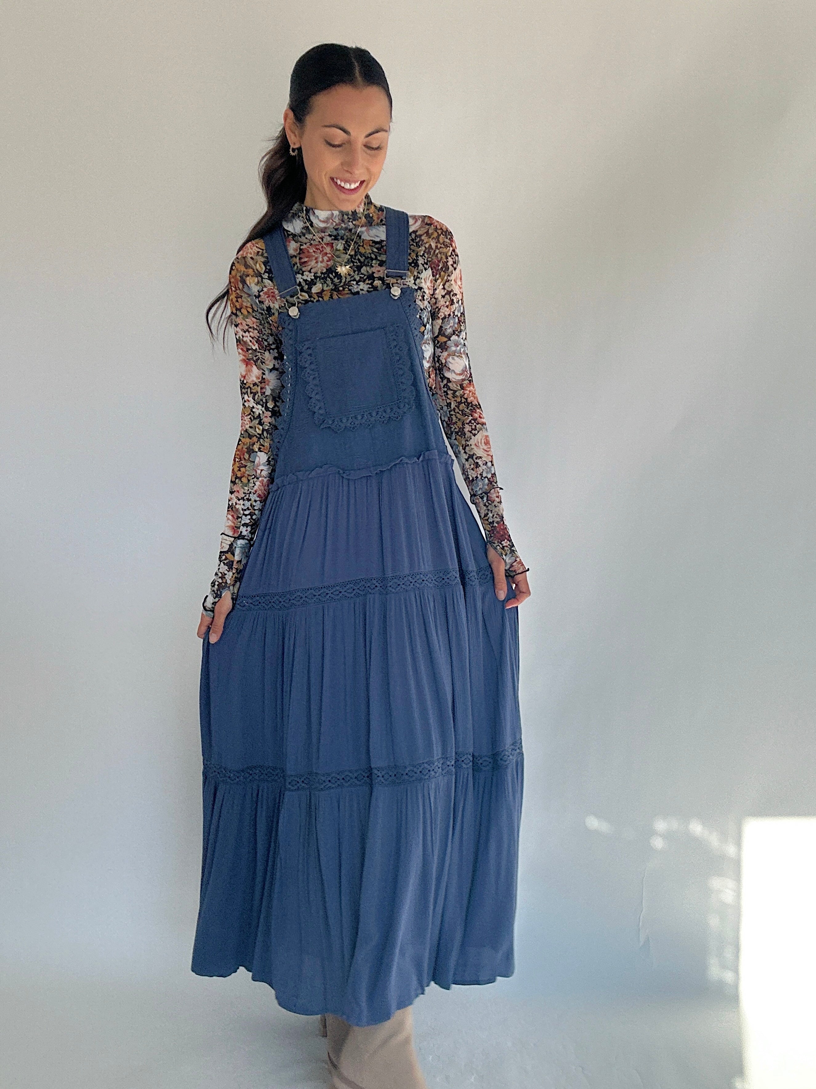 Woman wearing a blue overall dress with a floral long-sleeve shirt underneath against a white background