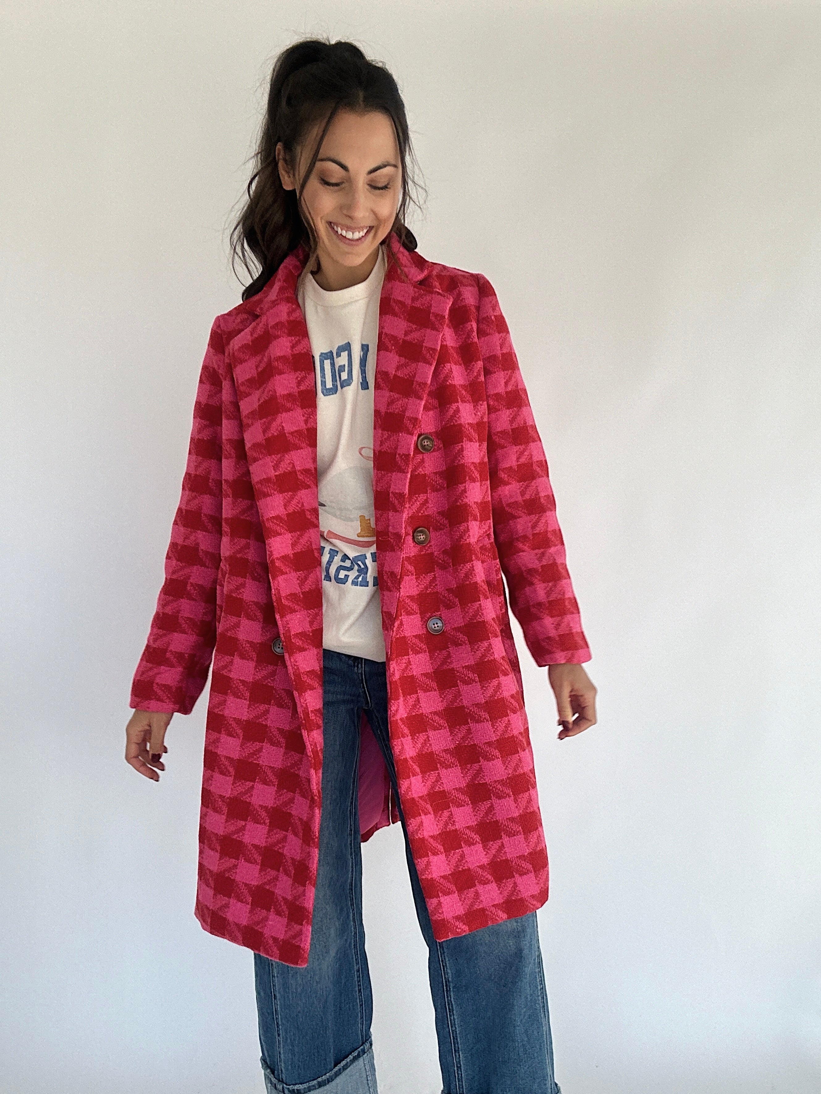 A person standing and smiling, wearing a hot pink and red houndstooth tweed coat with a double button front and lapel, paired with blue jeans.